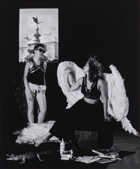 Black and white photograph. A young woman in oversized angel wings sits on a box with her head turned towards another woman with cropped hair, who wears sunglasses. Magazines, a crumpled sheet of plastic, and a toy alligator are spread across the floor, while a photograph of a city monument is projected on the wall. 