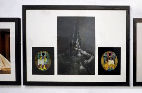 Installation photograph. Three framed artworks are hung closely together. The two on either side are cropped from view. The artwork in the centre is in three parts; a tall, central black and white photograph documents a dark toned man holding two large leaves over his face, while the smaller, flanking images are two colourful, oval designs.