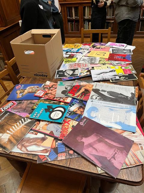 Photograph. A collection of colourful booklets and magazines are spread out across a brown wood tabletop.  Magazine titles include “Jock”, and “Zipper”. Also on the table is a brown cardboard box containing more material. The table has wood chairs pushed in against it, and three people stand beyond it.
