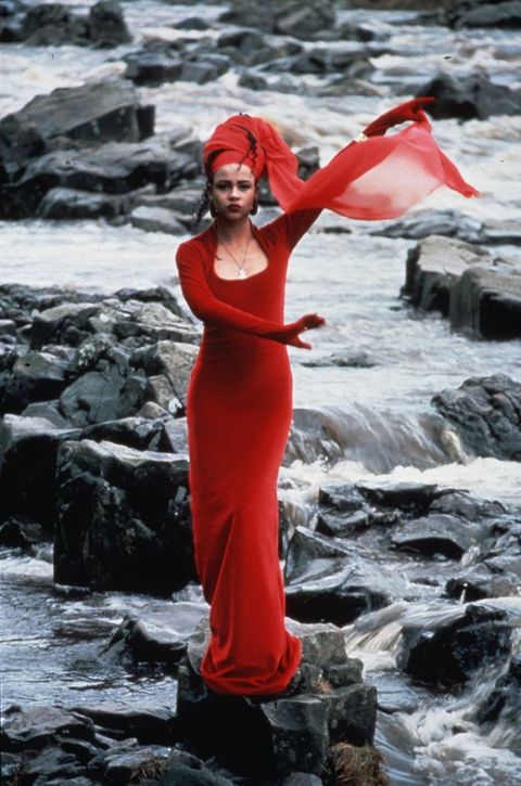 Colour film still. Woman with medium-brown skin tone wearing a figure-hugging long dress, her left hand extended upwards holding the end of her red voile headscarf. She stands centrally on rocks against a series of cascading water on dark rugged river rocks.