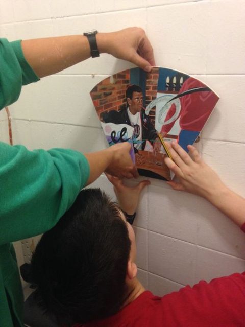 Colour photograph. An adult and child install a fan-shaped painting of a man with medium-brown skin seated in a café, on a white wall. The child holds a pencil as they adjust the artwork into place.