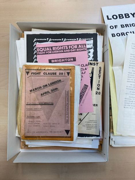 Digital Facsimile. A box contains a series of printed flyers and posters printed onto various sizes of white, pink, and yellow paper. One passage of visible text reads, “EQUAL RIGHTS FOR ALL, FIGHT FOR LEBIAN AND GAY RIGHTS”, while another flyer reads, “FIGHT CLAUSE 28!”. 