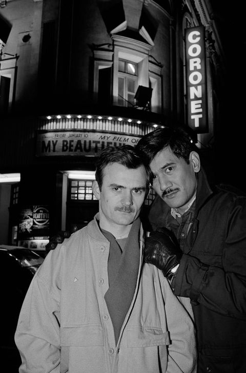 Black and white photograph. Print of two men standing, shown from the waist up, both wearing coats and one wearing leather driving gloves. In the background is an illuminated theatre building with the vertical signand text "CORONET" shown against a night sky.