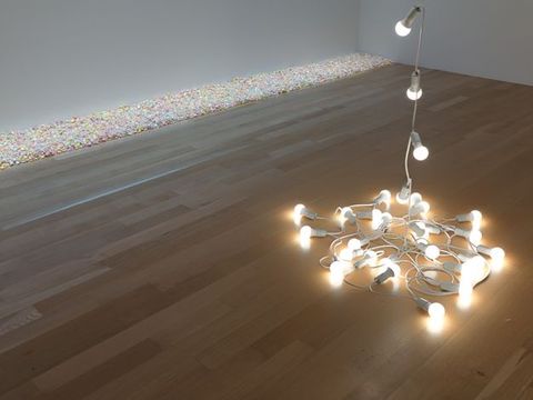 Installation. A string of globe lights in white casings pool on a pale wooden floor with a length suspended above the view of this interior space. A channel of irridescent pebbles arranged along one wall intersecting with another wall, in this corner view of a room.
