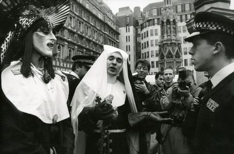 Black and white photograph. Two people wearing nun costumes stand before a policeman on a busy city street. One is wearing drag make-up and an elaborate headdress, and the other heavy leather gloves. A crowd of people gather behind them, one of whom is taking a photograph, while another carries a camera. City buildings are seen in the street beyond.