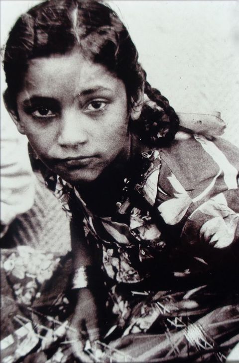 Black and white photograph. A south Asian child wearing bangles and a floral dress kneels on the floor, her head in the upper-left corner. She looks at the camera with sad eyes, her hands holding what appears to be wires.