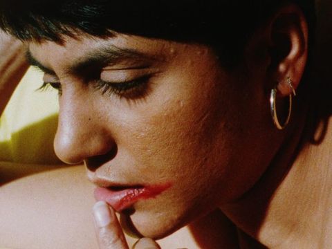 Colour film still. A naked woman wearing metallic gold earrings with short black hair gazes downwards, her right arm supporting her head. A finger touches the inside of her lip where red lipstick is smeared beyond the corner of her mouth, against a yellow background.