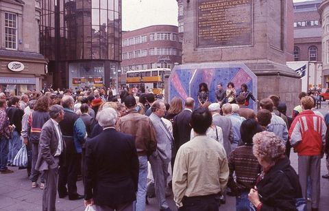 A black and white photograph of a performance of *The Last Blind Date Show* at Grey's Monument, Newcastle upon Tyne, 22 May 1992. The image shows a group of people gathered around a small stage, with a performer in the centre of the frame. The performer is wearing a white suit and is holding a microphone. The audience is visible in the background, with some people sitting on the ground and others standing. The monument is visible in the background, with a large crowd of people gathered around it. The sky is overcast, and the ground is wet, suggesting that it has recently rained.