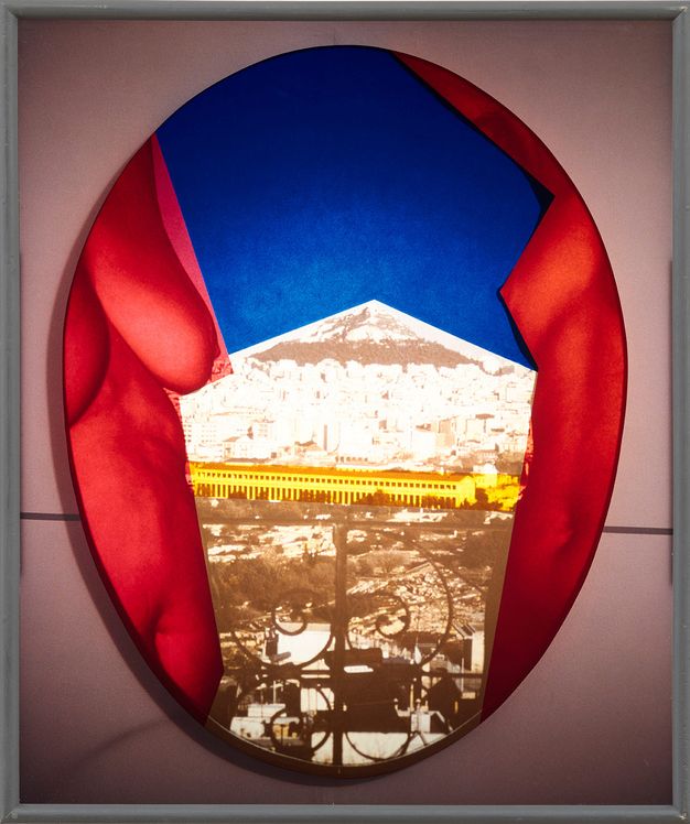 Installation. Within an oval shape, the partially-seen torsos of two women are rendered in bright red. Between them is a flat, dark blue shape, below which is a fragmented, sepia-tinted photographic image showing elements of a city. Running through the photograph image is a bright yellow stripe. 