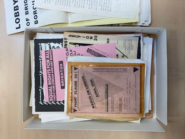 Digital Facsimile. A box contains a series of printed flyers and posters printed onto various sizes of white, pink, and yellow paper. One passage of visible text reads, “EQUAL RIGHTS FOR ALL, FIGHT FOR LEBIAN AND GAY RIGHTS”, while another flyer reads, “FIGHT CLAUSE 28!”. 