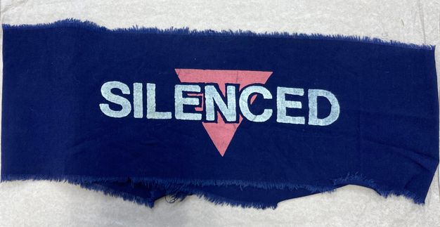 Colour photograph. This colour photograph documents a roughly-cut armband made from thin blue fabric. It features a printed logo made from a pink triangle, overlaid with the white text reading, “SILENCED”. 